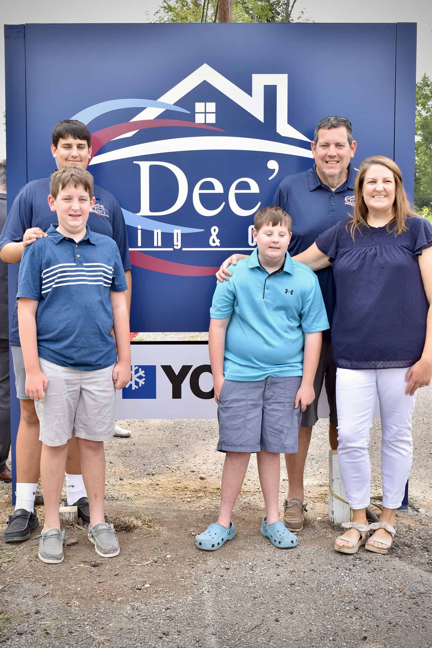 A family of five—two adults and three boys—stand smiling in front of a large blue Dees Heating & Cooling sign outdoors, dressed casually in blue and grey clothing.
