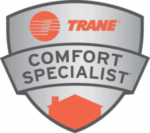 A silver badge with the orange Trane logo and the words TRANE COMFORT SPECIALIST in black text, with an orange silhouette of a house at the bottom.