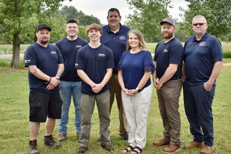 A group of seven adults, five men and two women, stand outdoors on grass, smiling at the camera. They are dressed casually, mostly wearing navy blue shirts, with trees and greenery in the background.