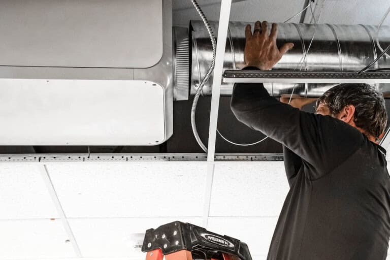 A worker stands on a ladder and installs or repairs an air duct in a ceiling, with ceiling tiles removed to access the ventilation system.