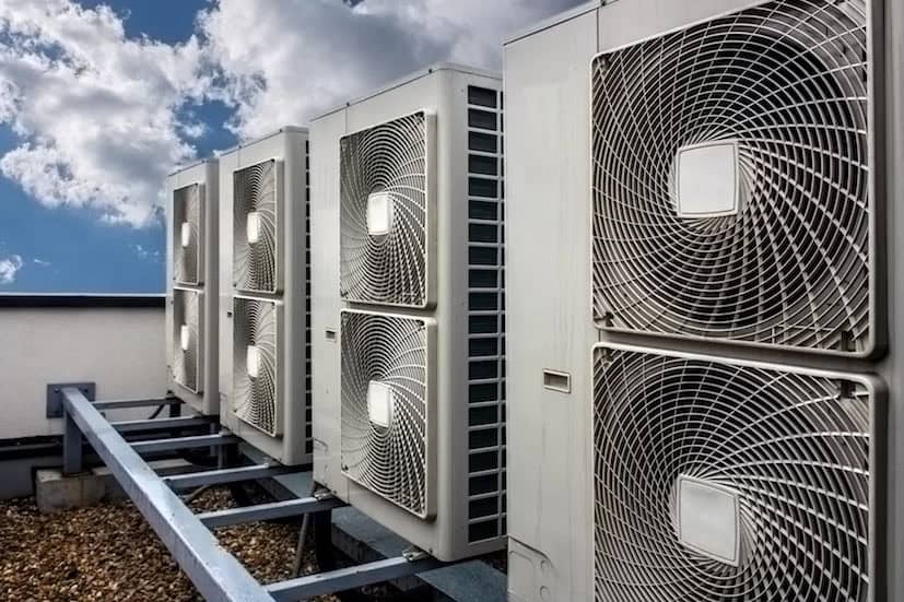 Large industrial air conditioning units are mounted outdoors on a rooftop platform under a partly cloudy sky. The metal units feature prominent circular fan grilles.