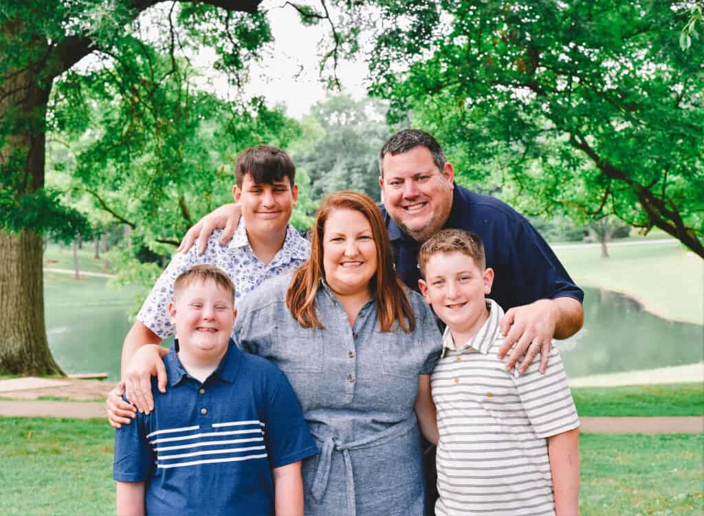 A smiling family of five poses outdoors in a park near a pond, surrounded by green trees. Two boys stand in front, with another boy and their parents standing behind them, all looking at the camera.