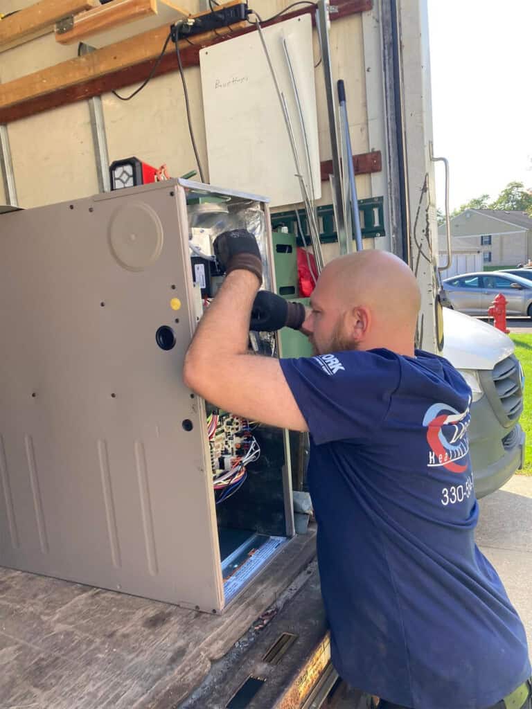 A technician in a navy shirt repairs or inspects a large metal HVAC unit on the back of a truck, using a power drill. Tools and equipment are visible around him, and a house can be seen in the background.