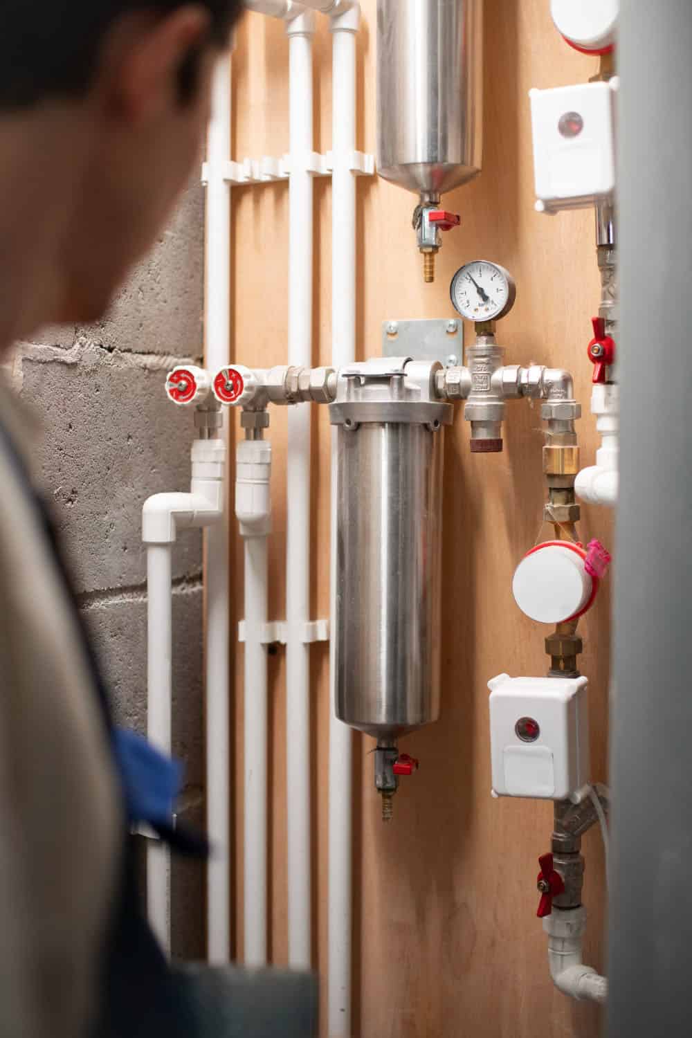 A person observes a wall-mounted plumbing system with pipes, valves, a pressure gauge, and filtration canisters in a utility room.