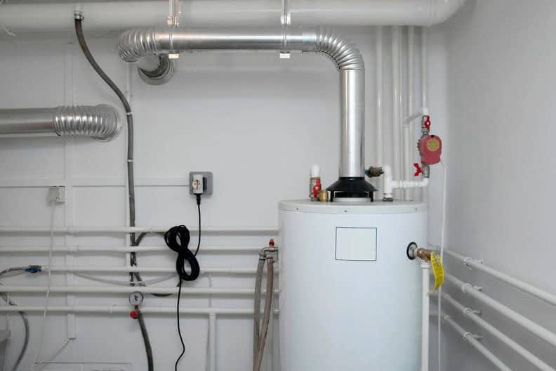 A white water heater with metal pipes and ventilation ducts attached, installed in a clean, white-walled utility room. Various pipes run along the walls, and a power outlet is visible.