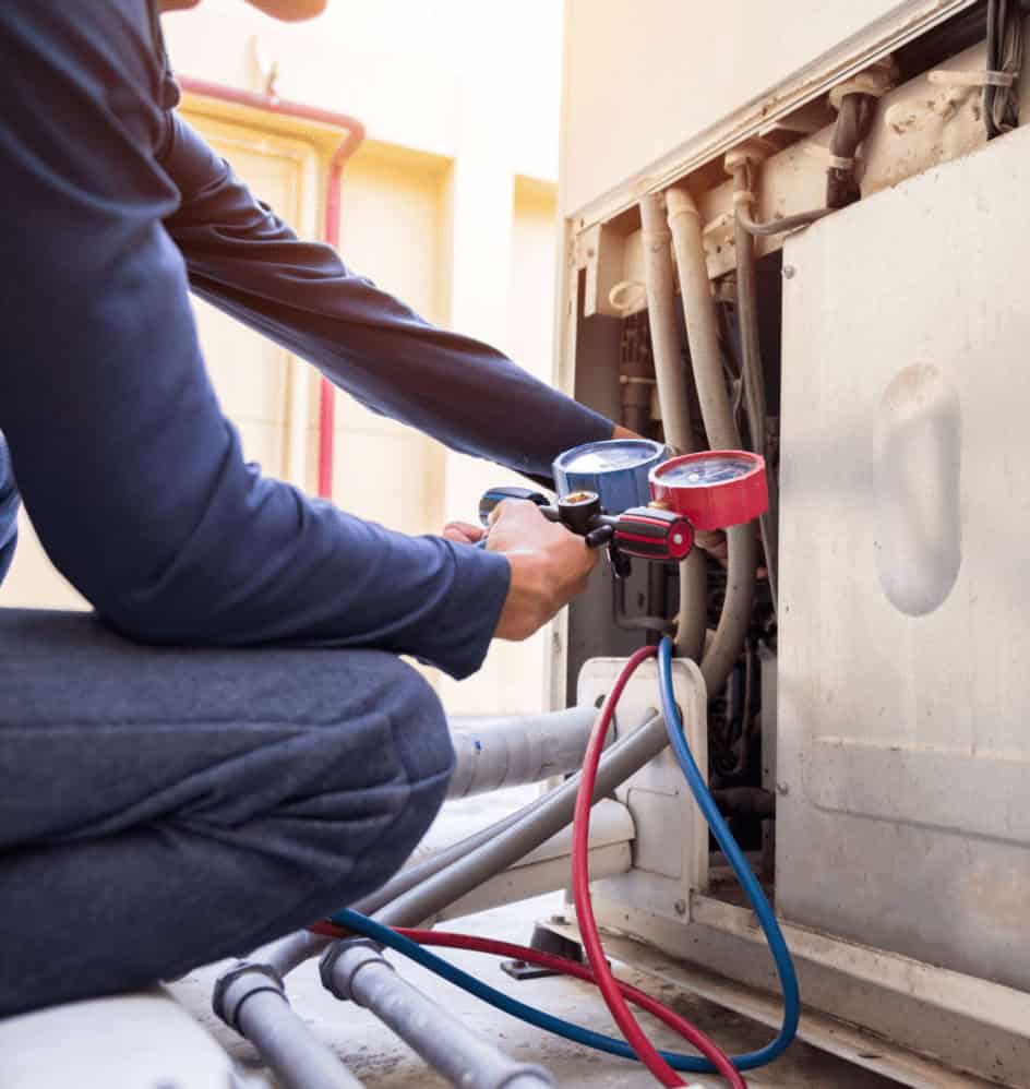 A technician in blue workwear uses a pressure gauge and colored hoses to inspect or repair an outdoor HVAC unit. The technician is crouched beside the equipment, checking connections.