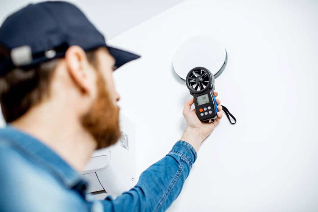 A person wearing a blue cap and denim shirt uses a handheld anemometer to measure airflow near a round vent on a white wall. The device’s display screen is visible.