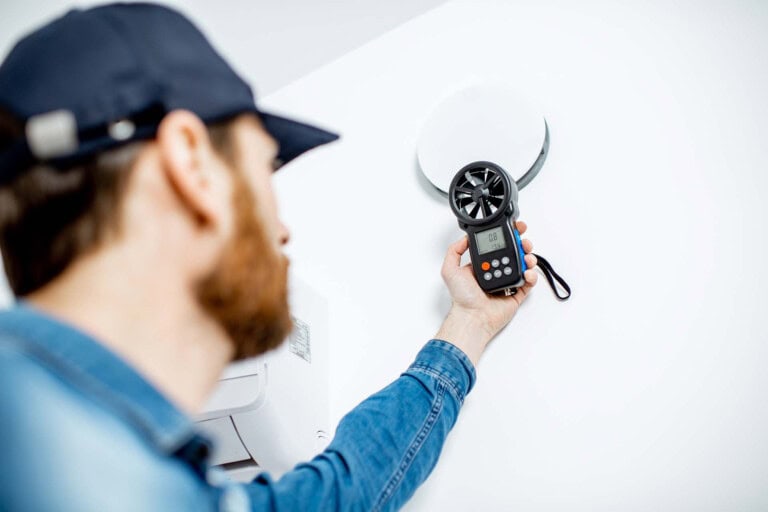 A person wearing a blue cap and denim shirt uses a handheld anemometer to measure airflow near a round vent on a white wall. The device’s display screen is visible.