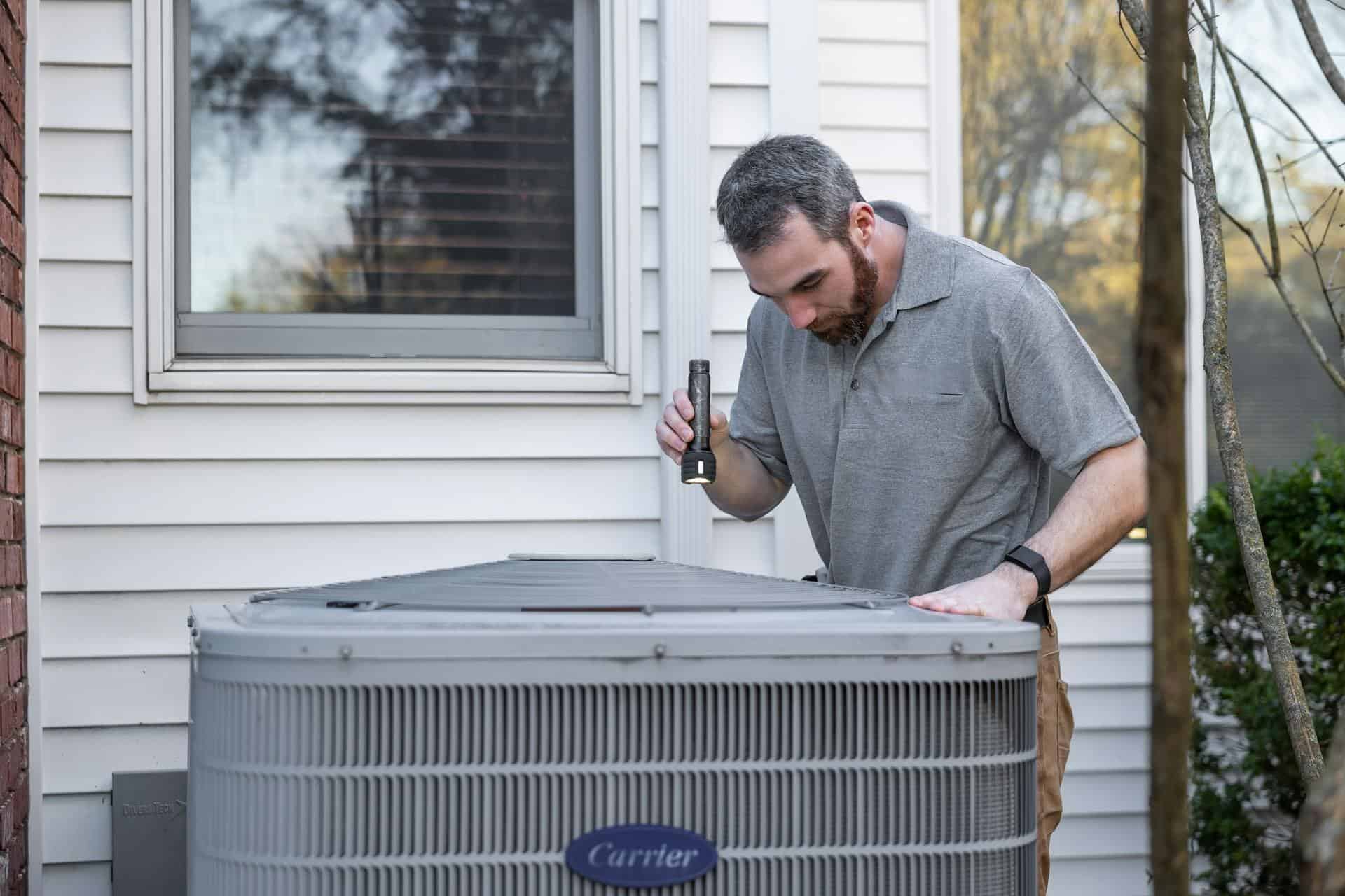 A man inspects an outdoor air conditioning unit with a flashlight next to a house with white siding and a window.