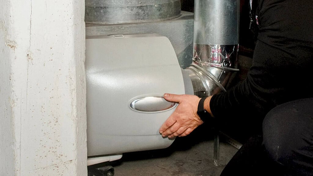 A person installs or adjusts a humidifier unit attached to the ductwork of a home HVAC system, with their hands on the metal and plastic components.