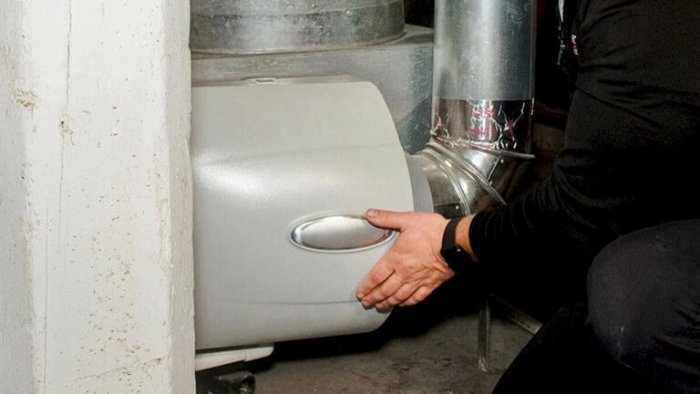 A person installs or adjusts a humidifier unit attached to the ductwork of a home HVAC system, with their hands on the metal and plastic components.