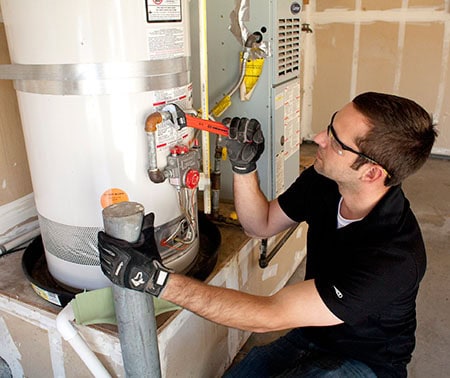 A man wearing safety glasses and gloves uses a wrench to adjust a valve on a water heater in a garage or utility room. A furnace and unfinished drywall are visible in the background.