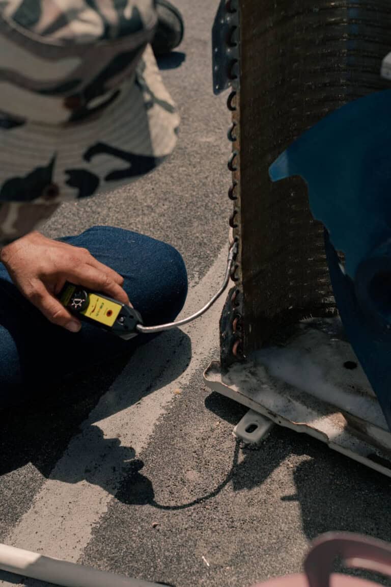 A person uses a yellow digital multimeter to check the wiring or electrical connections of a metal appliance or HVAC unit on a textured outdoor surface.