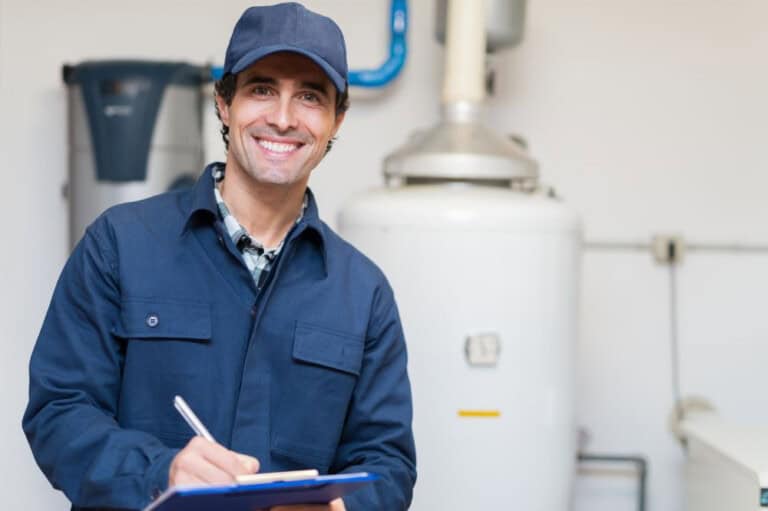 Smiling maintenance worker in a blue uniform and cap holds a clipboard and pen, standing in front of a water heater and pipes in a utility room.