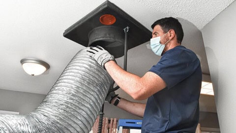 A man wearing a face mask and gloves is cleaning an air duct in a ceiling with a large flexible hose and specialized equipment in a home interior.