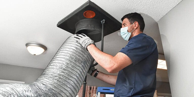 A man wearing a face mask and gloves is cleaning an air duct in a ceiling with a large flexible hose and specialized equipment in a home interior.