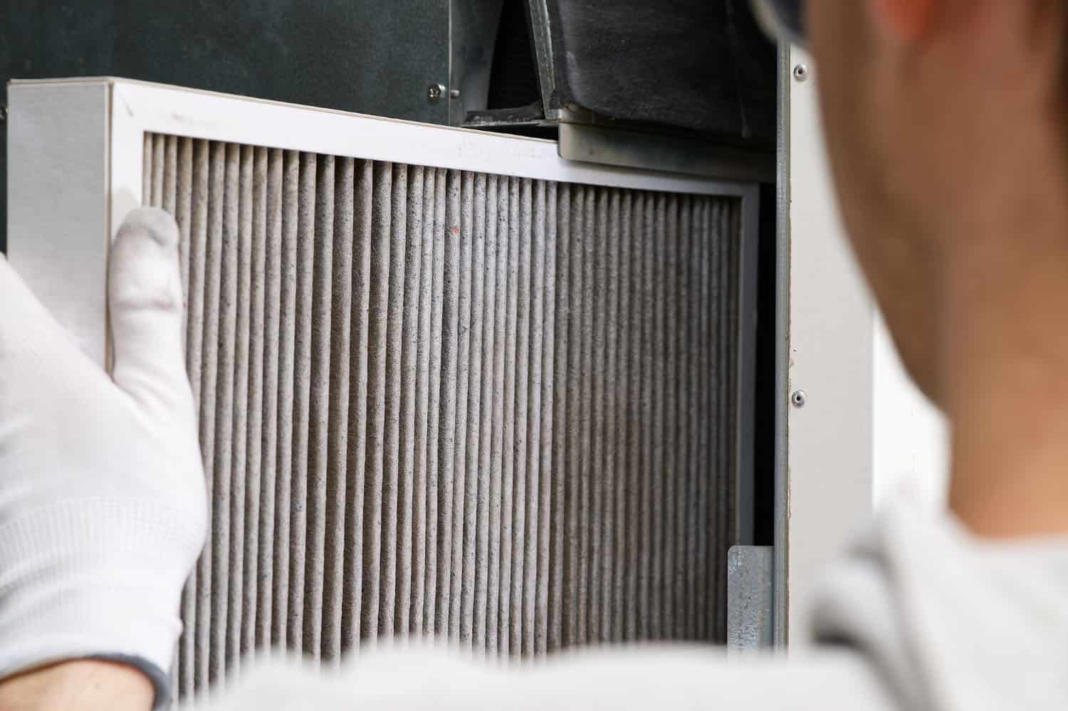A person wearing white gloves is installing or removing a dirty air filter from a ventilation or HVAC system. The filter appears to have collected dust and debris.
