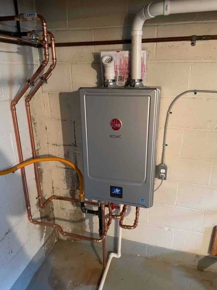 A wall-mounted Rheem IKONIC tankless water heater is installed in a basement, with copper pipes, a yellow gas line, and white PVC vent pipes connected. The wall is bare concrete blocks.