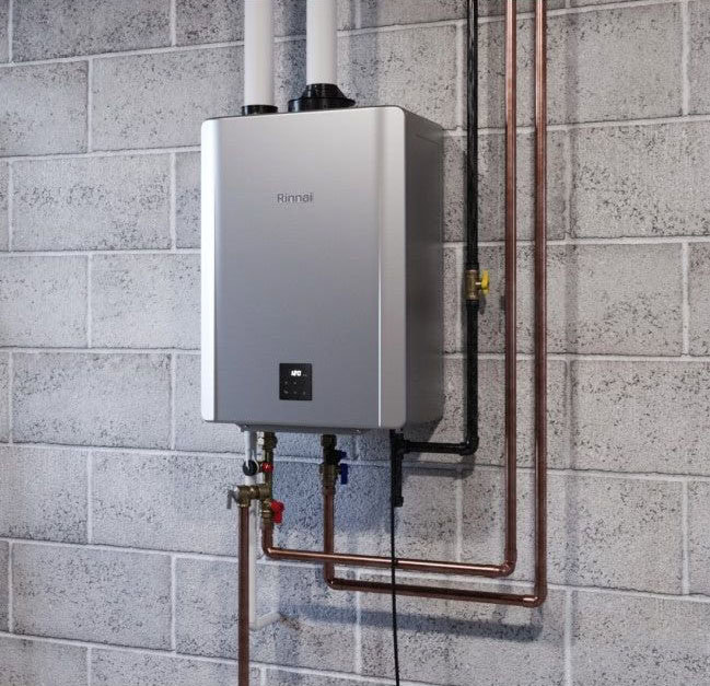 A tankless water heater with copper pipes mounted on a gray cinder block wall, featuring two white exhaust pipes extending upward.