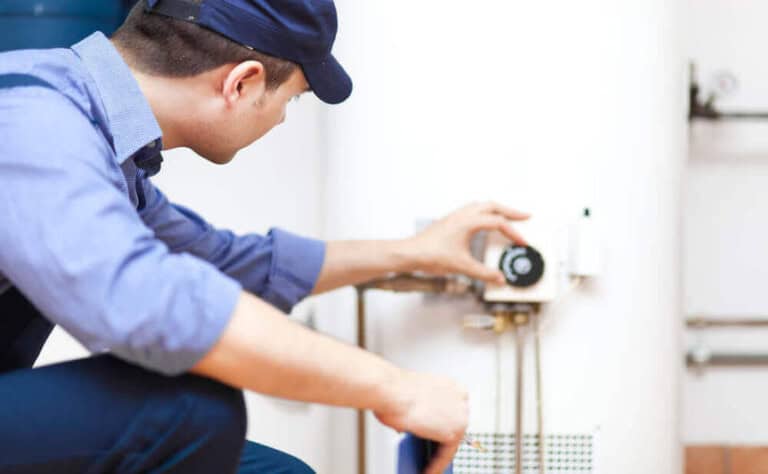 A plumber in a blue uniform and cap inspects and adjusts the control panel of a water heater in a utility room.