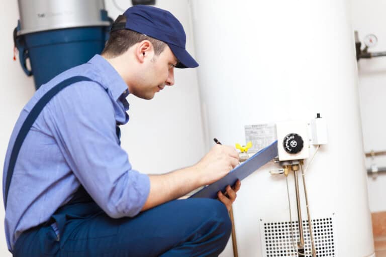 A man in work uniform and cap kneels beside a water heater, inspecting it and writing notes on a clipboard.