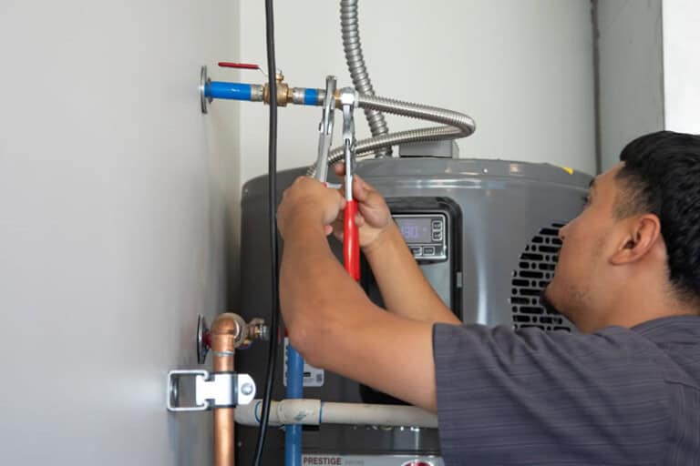 A person uses a wrench to tighten a pipe fitting on a water heater mounted against a wall, performing maintenance or installation work.