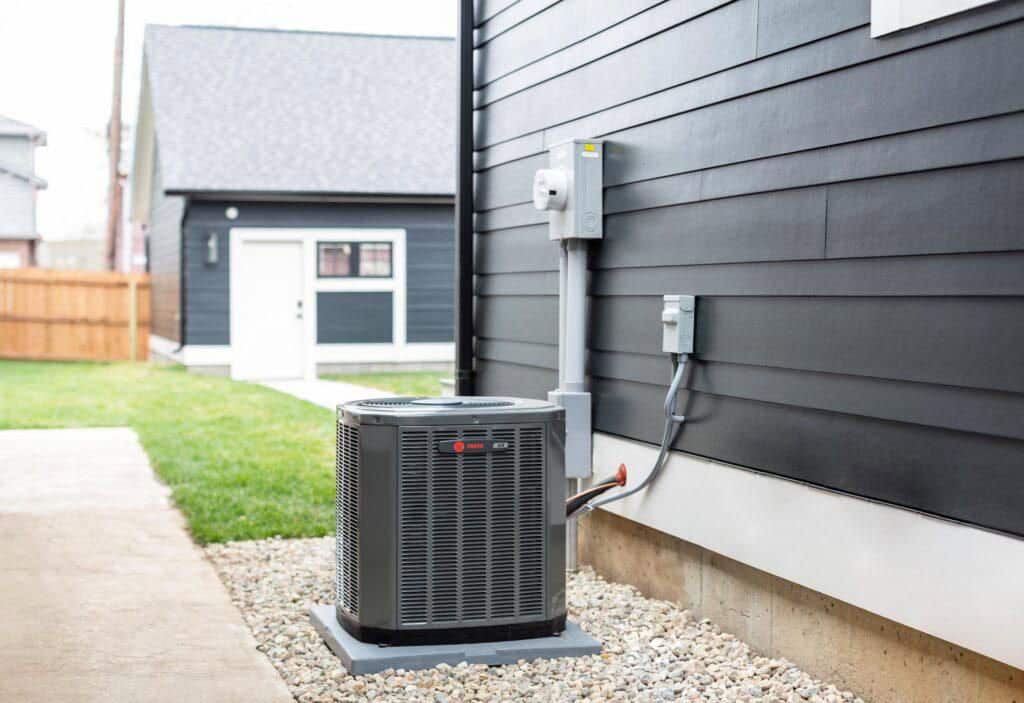 A central air conditioning unit sits on a concrete pad next to a dark gray house, surrounded by gravel and connected to electrical boxes on the exterior wall. A lawn and detached garage are visible in the background.