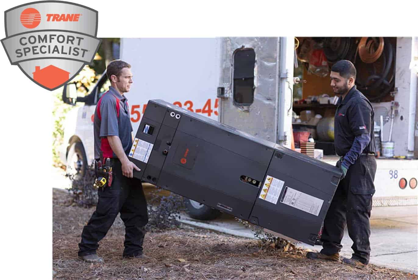 Two technicians move a large HVAC unit from a truck. Both wear black uniforms with red accents. The Trane Comfort Specialist logo is in the top left corner of the image.