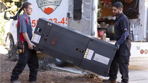 Two technicians in uniforms carry a large black HVAC unit outside, next to a white service van with tools and equipment in the open back.
