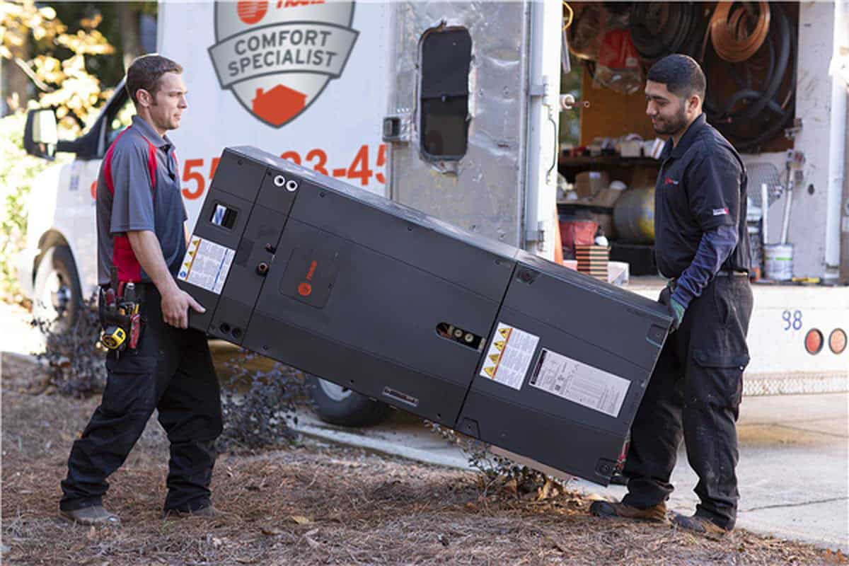Two technicians in uniforms carry a large black HVAC unit outside, next to a white service van with tools and equipment in the open back.