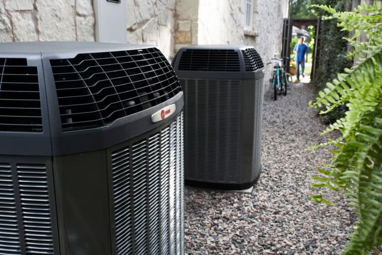 Two large outdoor air conditioning units sit on gravel beside a stone house; green plants are visible on the right, and a person and bicycle appear in the background.