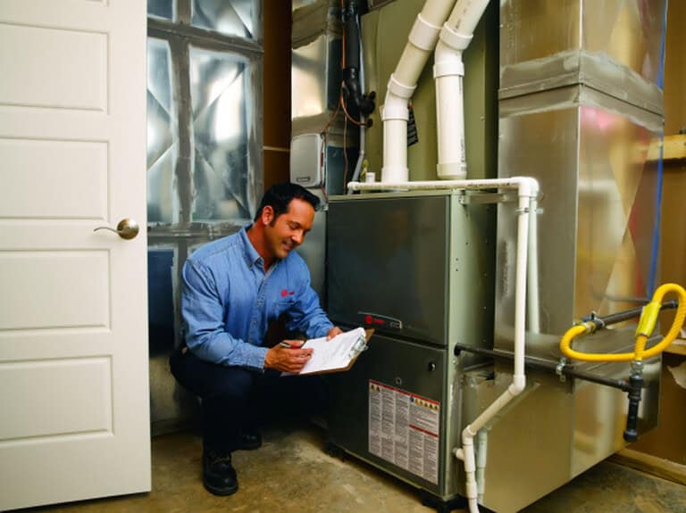 A technician in a blue shirt kneels next to a furnace in a utility room, smiling and writing on a clipboard while inspecting the HVAC system.