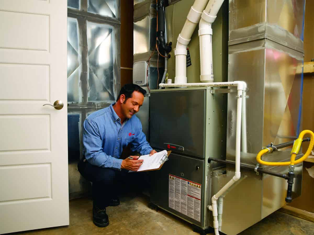 A technician in a blue shirt kneels next to a furnace in a utility room, smiling and writing on a clipboard while inspecting the HVAC system.